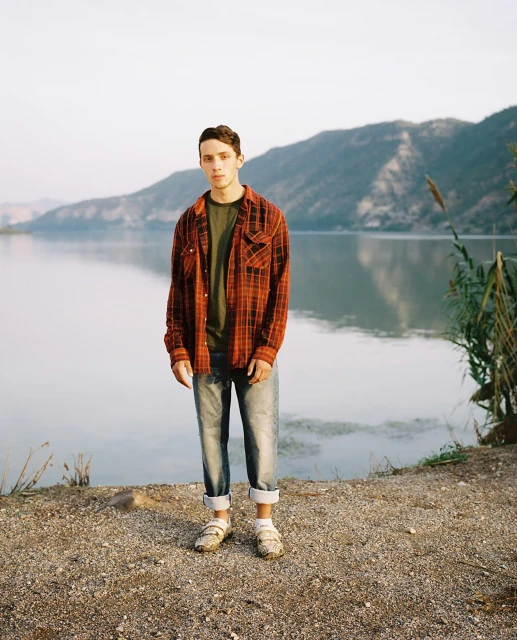 A young adult stands in a red plaid shirt and jeans near a lake with mountains in the background under a bright, overcast sky.
