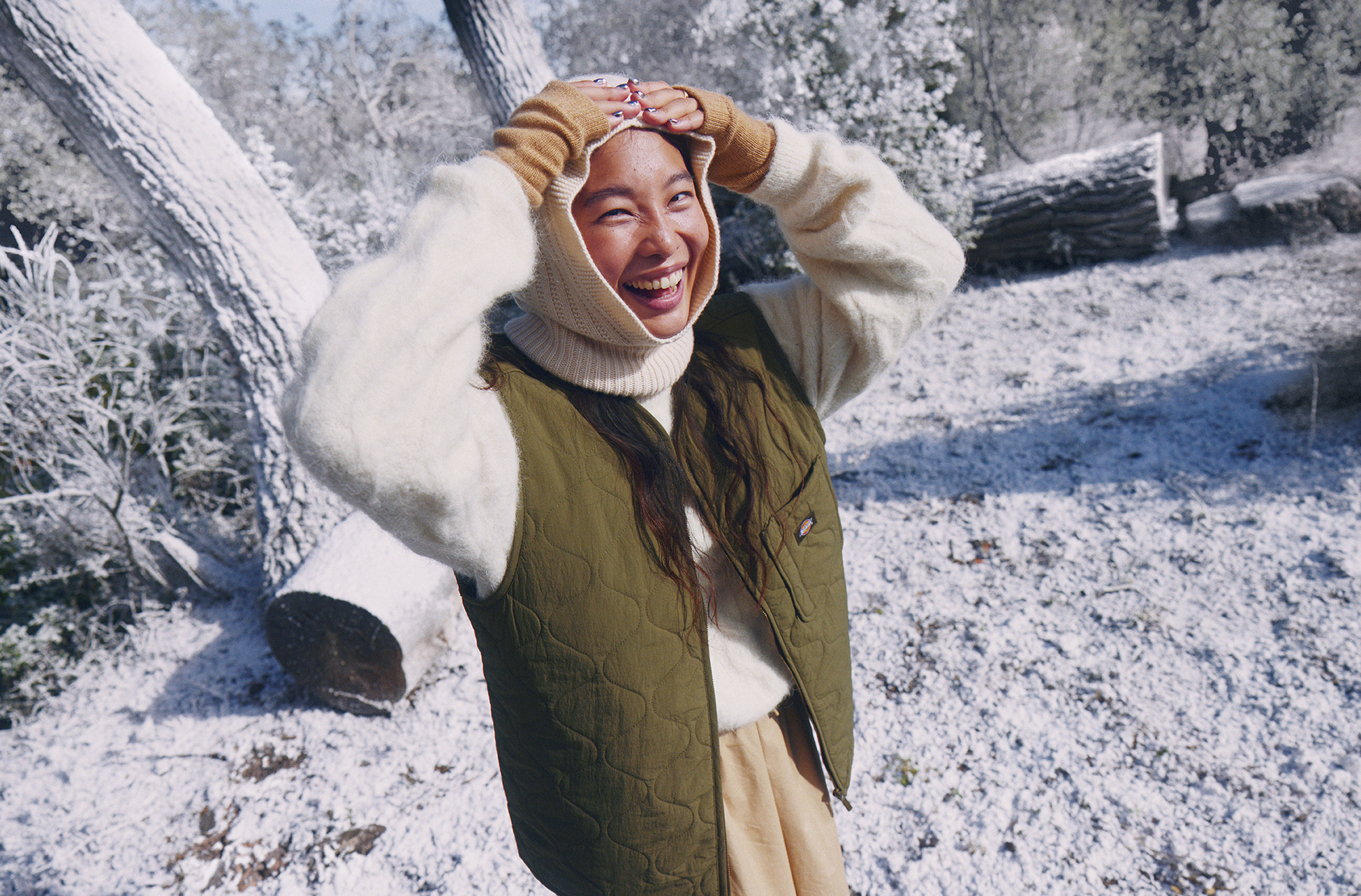 Smiling adult woman outdoors in winter apparel. She has her hands up with a snowy, woodland environment in the background.