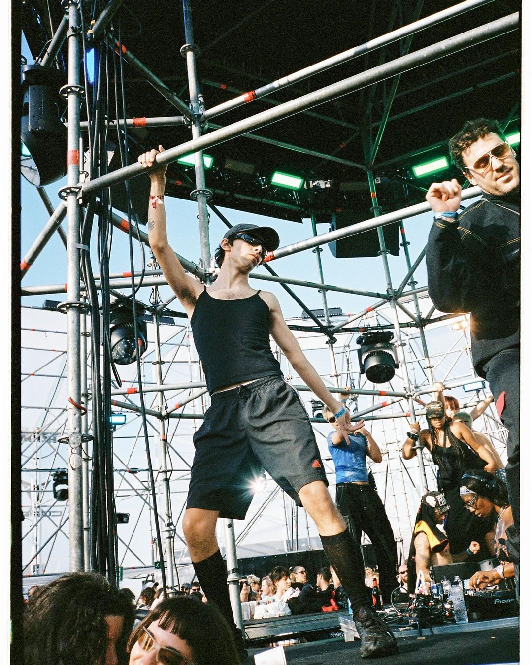Live concert scene featuring a performer on stage hanging on scaffolding with other people on stage and an audience in the foreground.