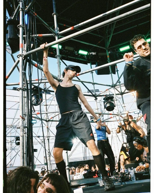 Live concert scene featuring a performer on stage hanging on scaffolding with other people on stage and an audience in the foreground.