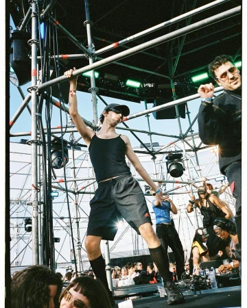 Live concert scene featuring a performer on stage hanging on scaffolding with other people on stage and an audience in the foreground.