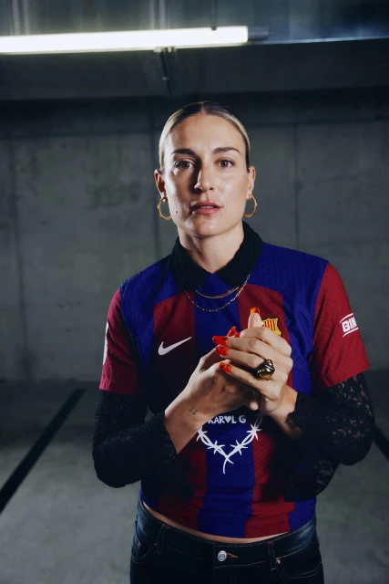 Woman in soccer jersey with red nails and gold jewelry in an indoor parking garage. Modern fashion meets sporty casual style.
