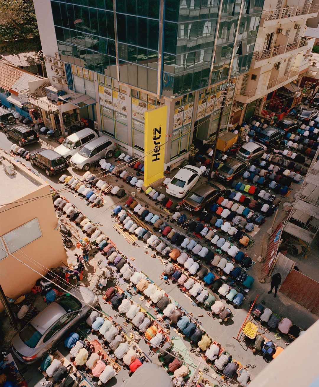 Aerial view showcases a large group of people kneeling in prayer on city streets, bordered by cars and a tall building with a Hertz sign.