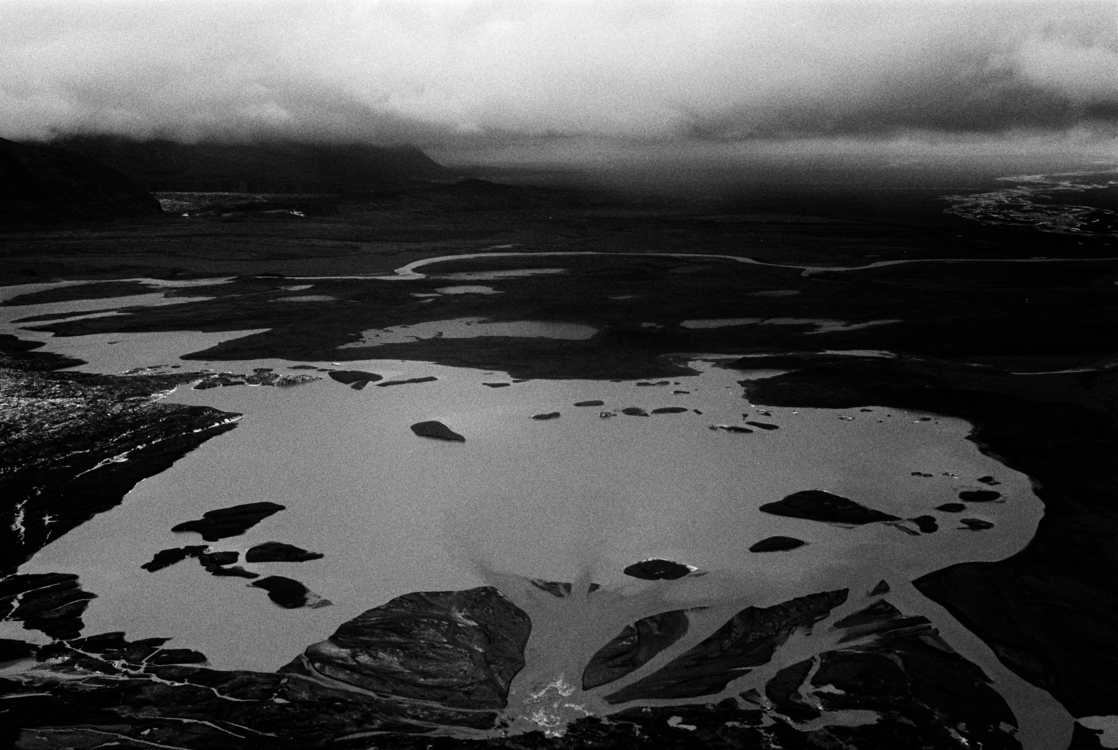 Monochrome aerial photograph of a glacial landscape with rivers, lakes, and dark, cloudy skies providing a stark, dramatic effect.