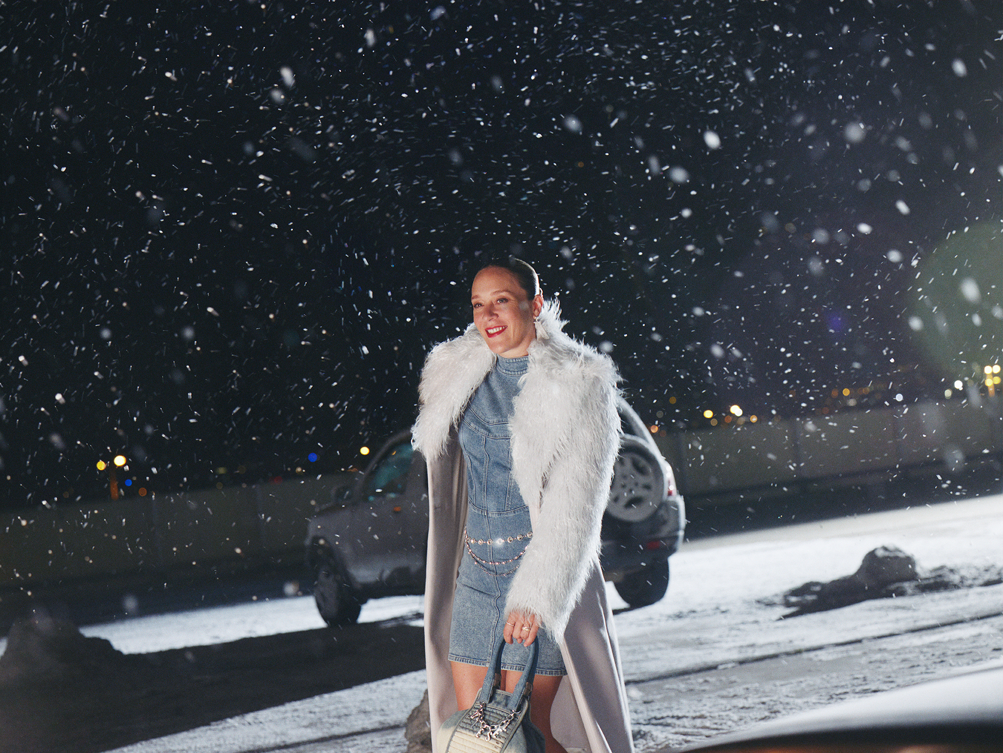 An adult woman stands outdoors at night in a denim outfit and faux fur coat, holding a purse. Snow falls around her, with cars in the background.