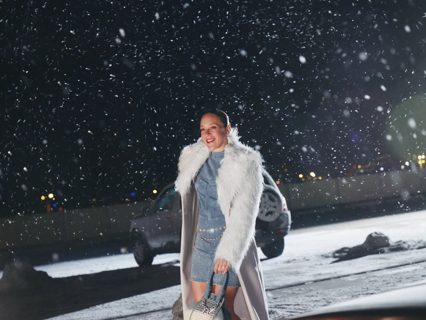 An adult woman stands outdoors at night in a denim outfit and faux fur coat, holding a purse. Snow falls around her, with cars in the background.