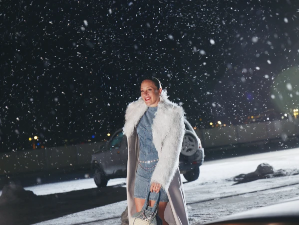 An adult woman stands outdoors at night in a denim outfit and faux fur coat, holding a purse. Snow falls around her, with cars in the background.