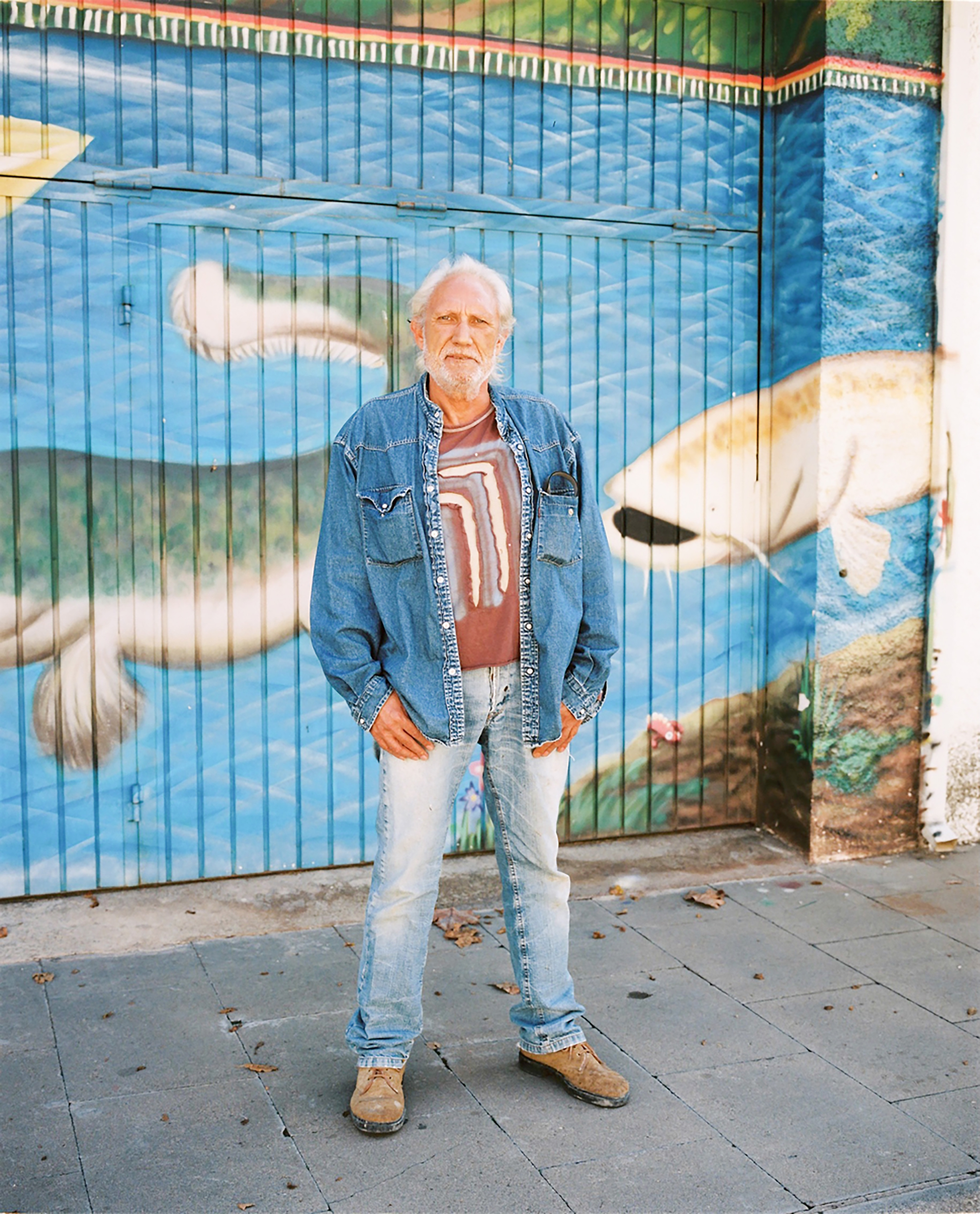 An elderly man with a white beard poses in front of a mural featuring fish, wearing a denim jacket, T-shirt, and light wash jeans.