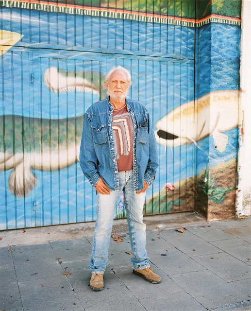 An elderly man with a white beard poses in front of a mural featuring fish, wearing a denim jacket, T-shirt, and light wash jeans.