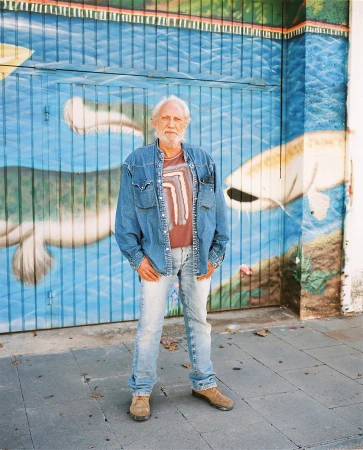An elderly man with a white beard poses in front of a mural featuring fish, wearing a denim jacket, T-shirt, and light wash jeans.