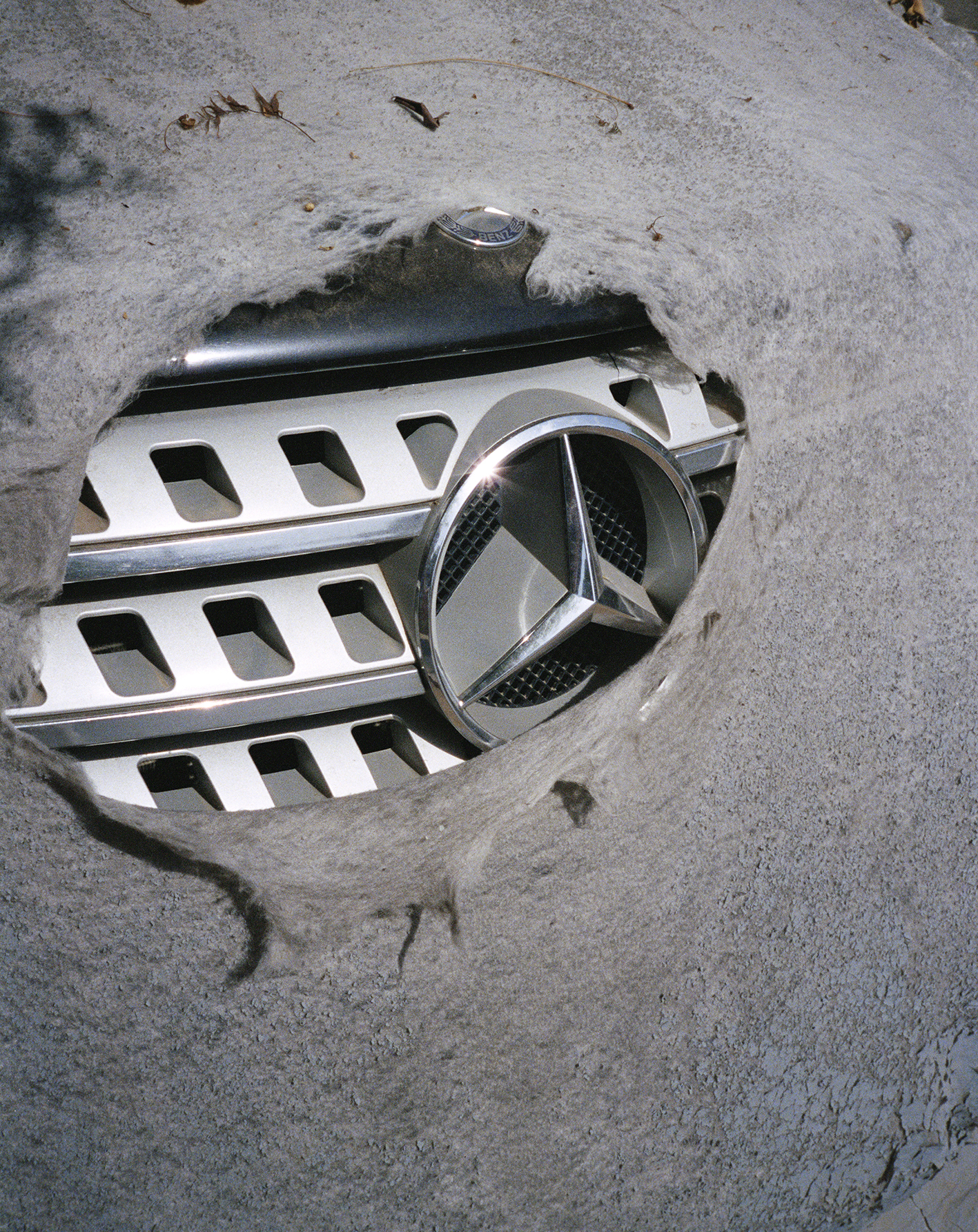 Mercedes-Benz logo and grill visible through a hole in a grey car cover. Close-up shot.