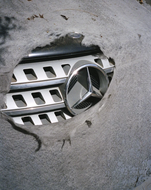 Mercedes-Benz logo and grill visible through a hole in a grey car cover. Close-up shot.
