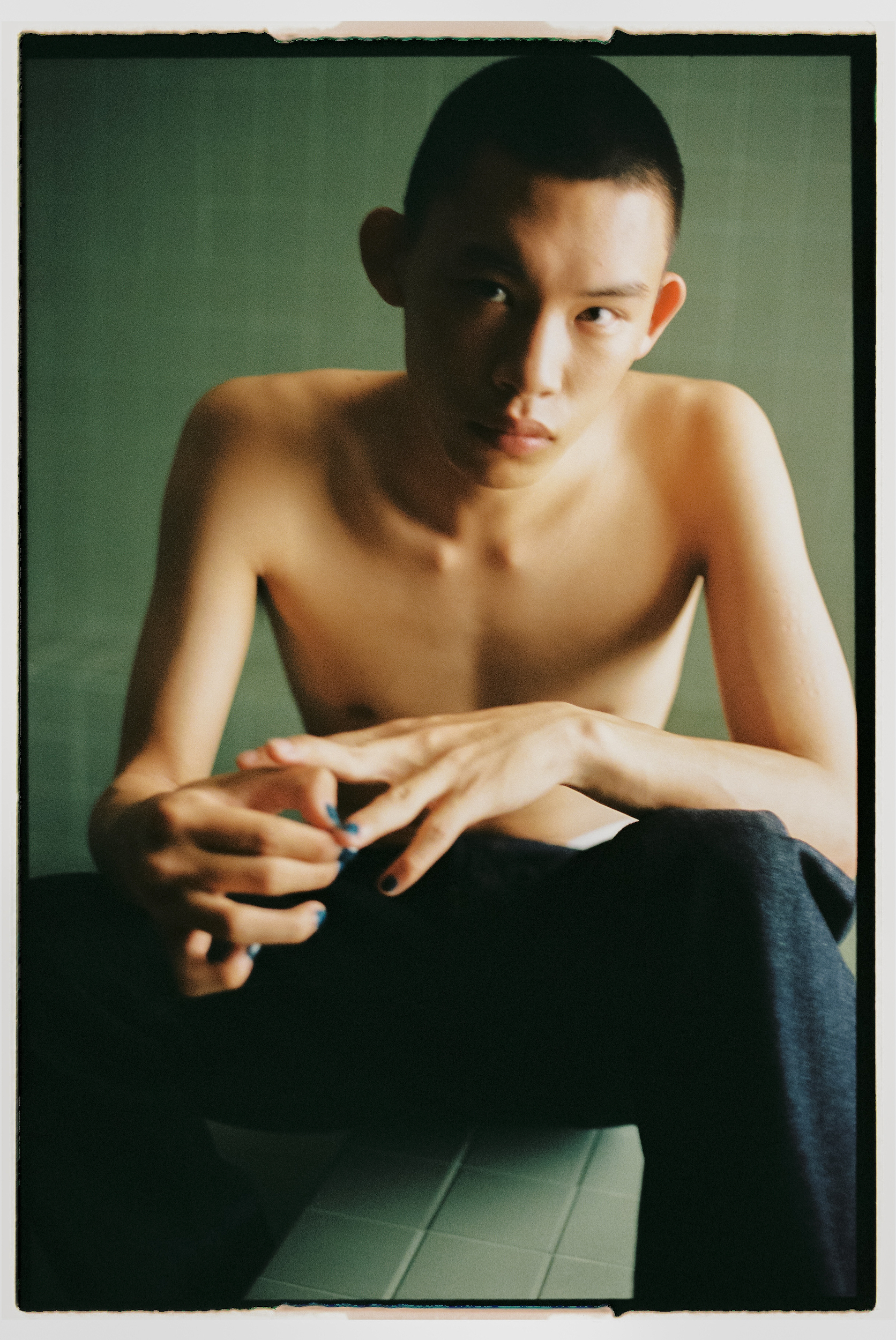 Portrait of a shirtless young East Asian man with short hair seated against a green tiled wall.