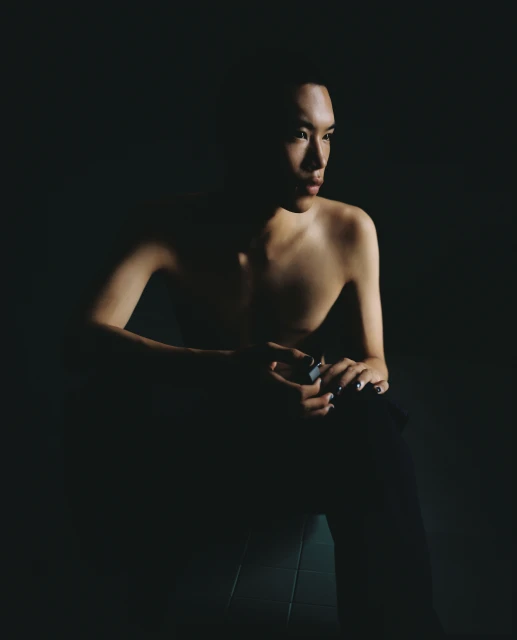 Portrait of a shirtless person with dark hair, seated in dark trousers against a dimly lit background with an introspective gaze.