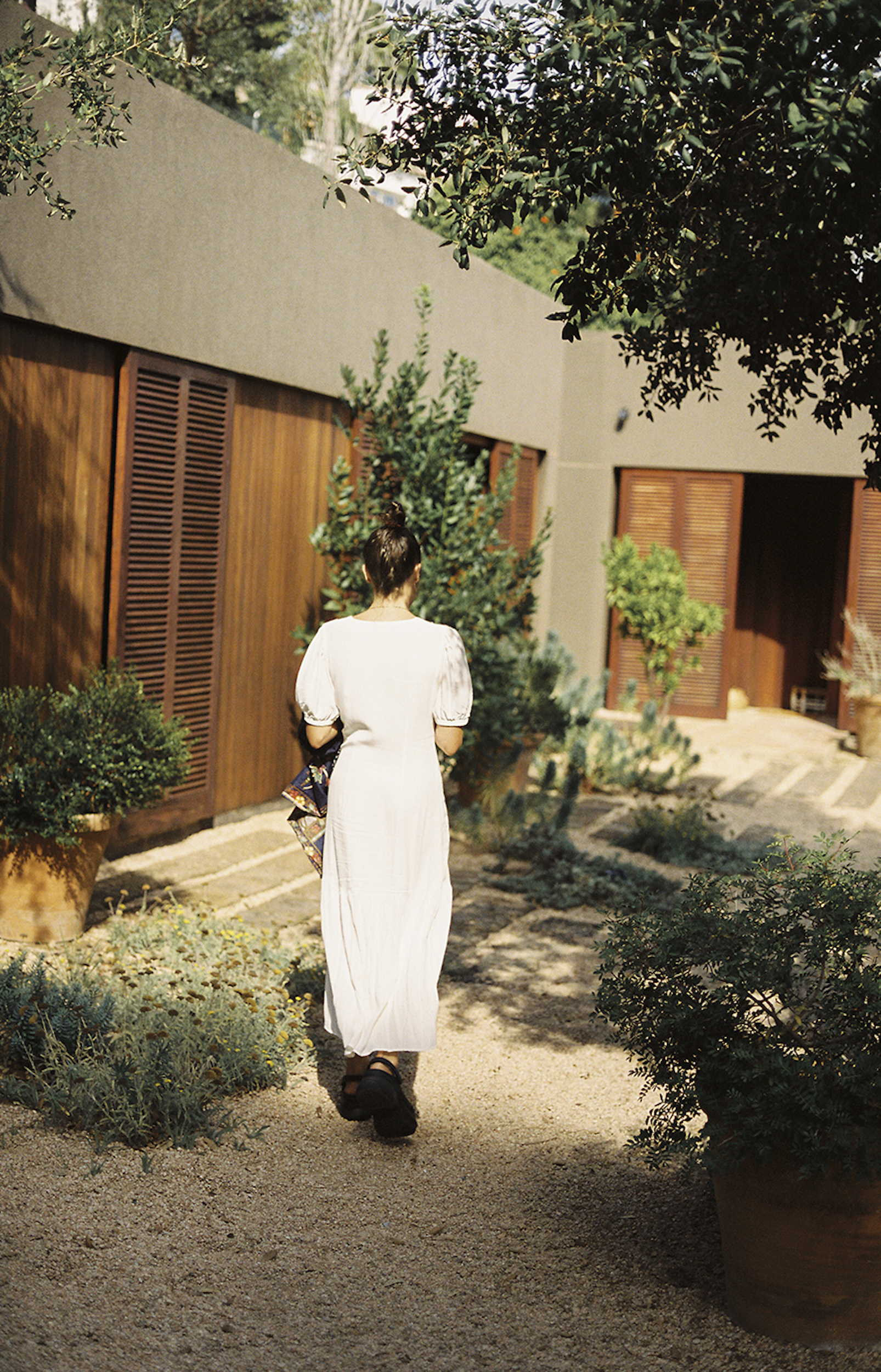 Woman in white dress walks along a gravel path surrounded by potted plants, with a modern building and lush greenery in the background.