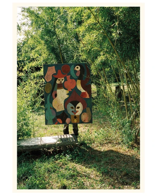 Person holding a colorful abstract painting in a green outdoor setting with bamboo, creating a juxtaposition of art and nature.