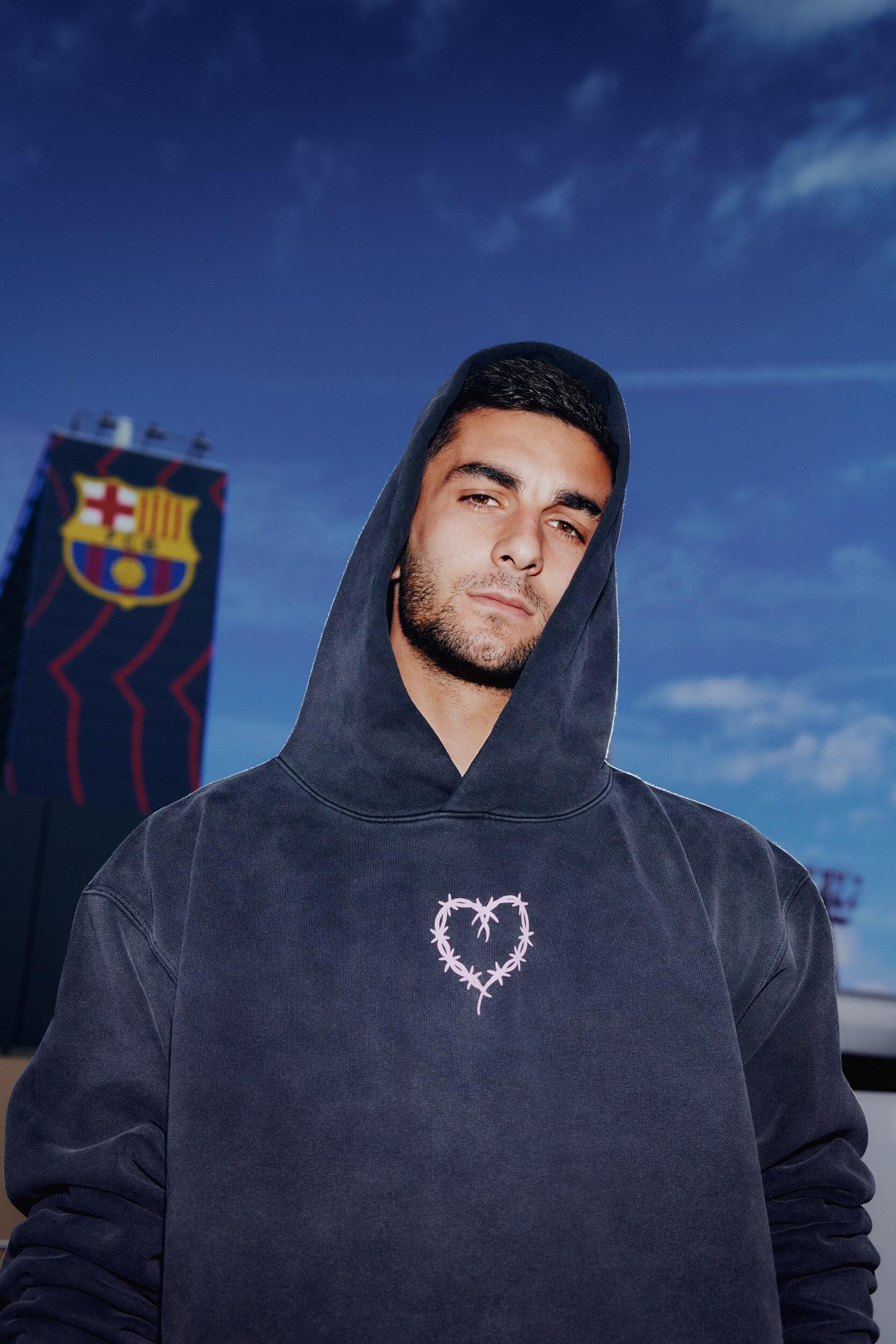 Young adult male in a hooded sweatshirt poses outdoors against a blue sky and a background building with a club emblem.