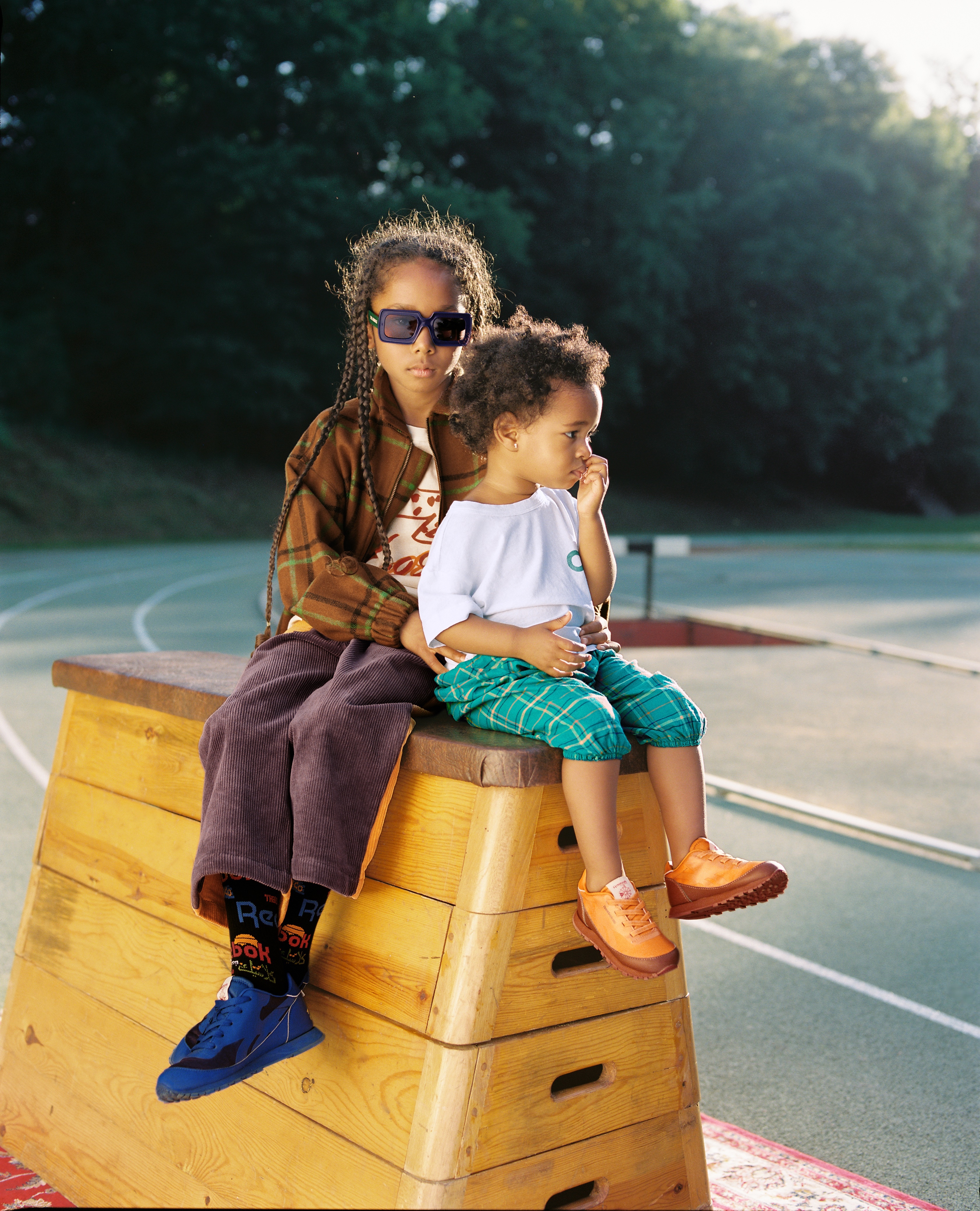 Two children sit on wooden gymnastic vault outdoors. One wears sunglasses, brown jacket, purple pants. Other wears shorts, orange shoes.