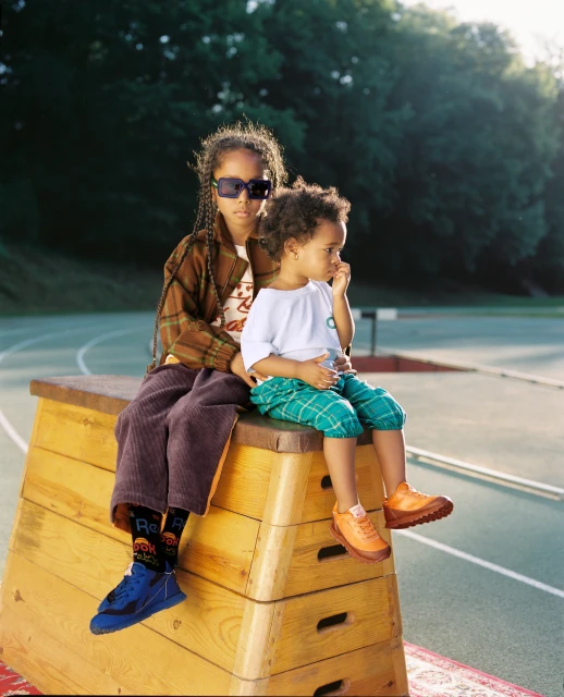 Two children sit on wooden gymnastic vault outdoors. One wears sunglasses, brown jacket, purple pants. Other wears shorts, orange shoes.