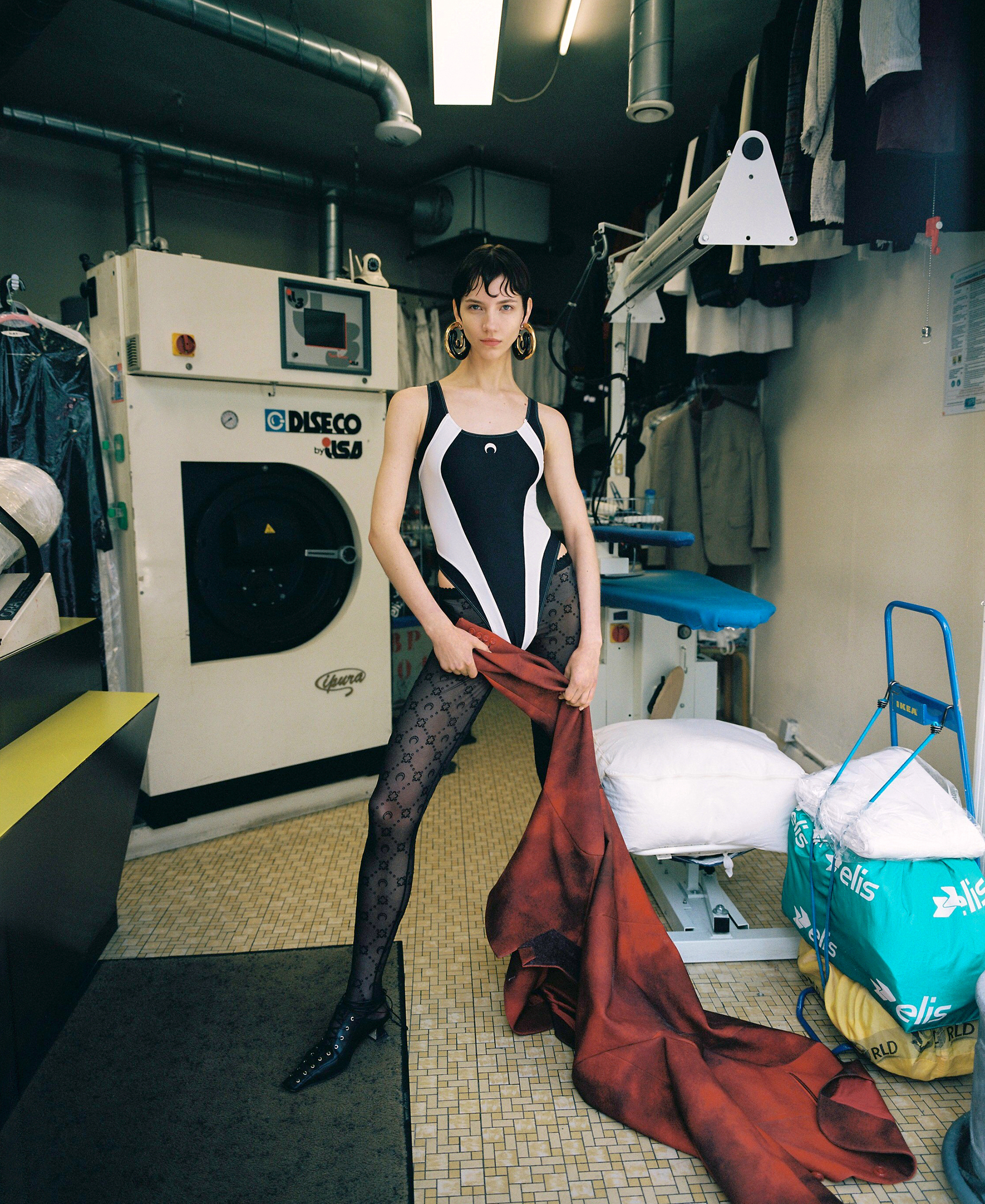 Woman in black and white bodysuit poses in laundry room with washing machine, ironing board, clothes, and patterned tights.