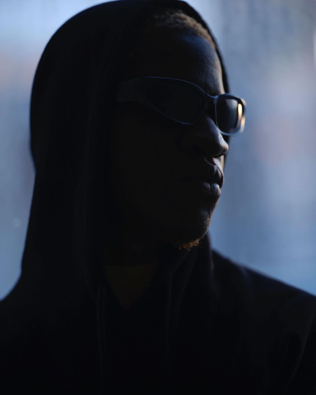 Portrait of African-American adult male with sunglasses and hoodie, facing to the right in front of a blurry blue background.