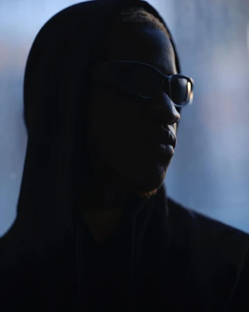 Portrait of African-American adult male with sunglasses and hoodie, facing to the right in front of a blurry blue background.