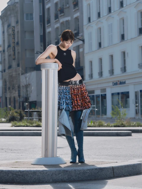 Young woman in modern fashion standing in urban plaza against city buildings in a sunlit, full outdoor shot with a silver column.
