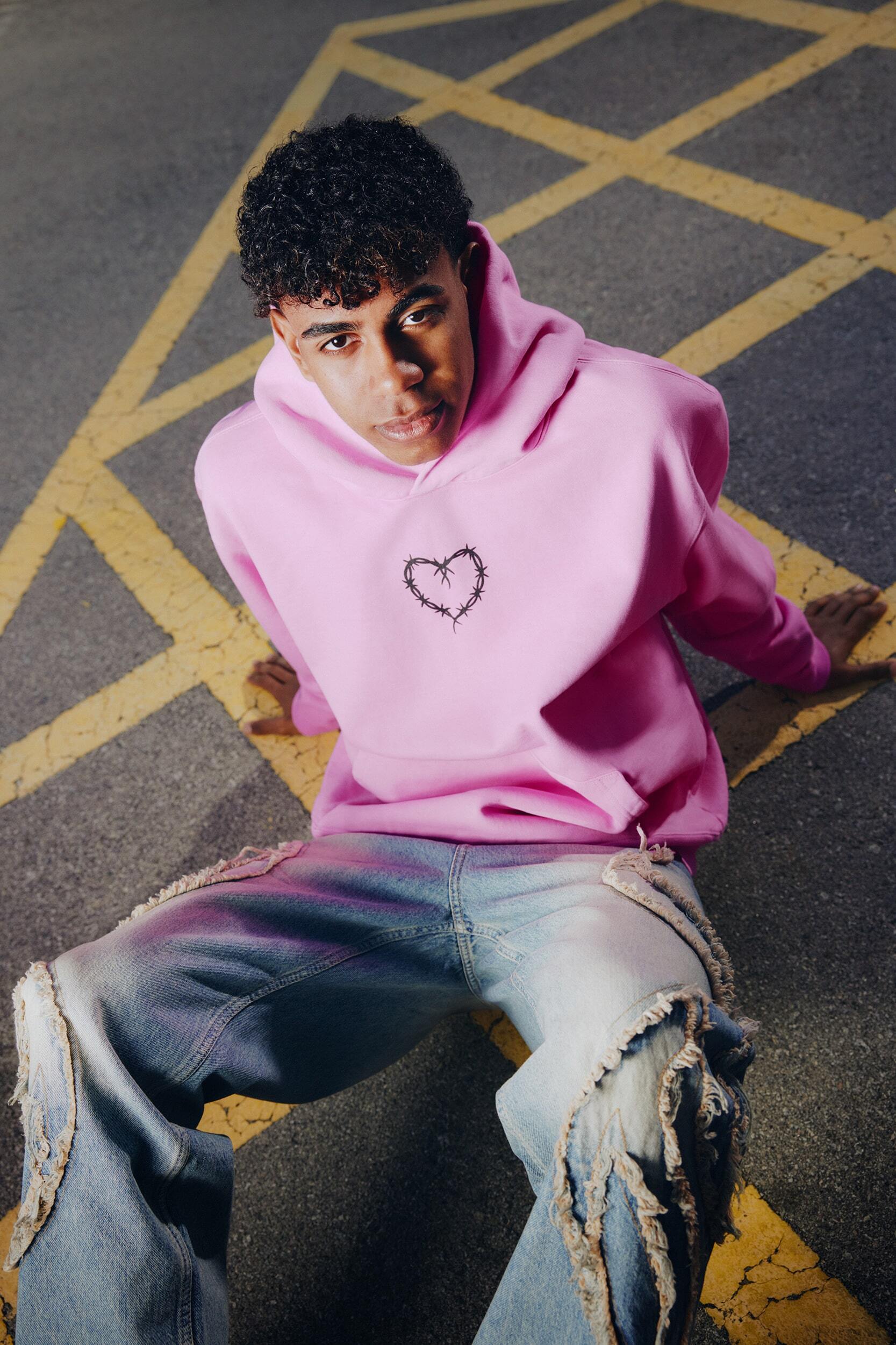 Young adult poses in pink hooded sweatshirt and distressed jeans against yellow-marked asphalt. Fashion-forward urban casual wear.