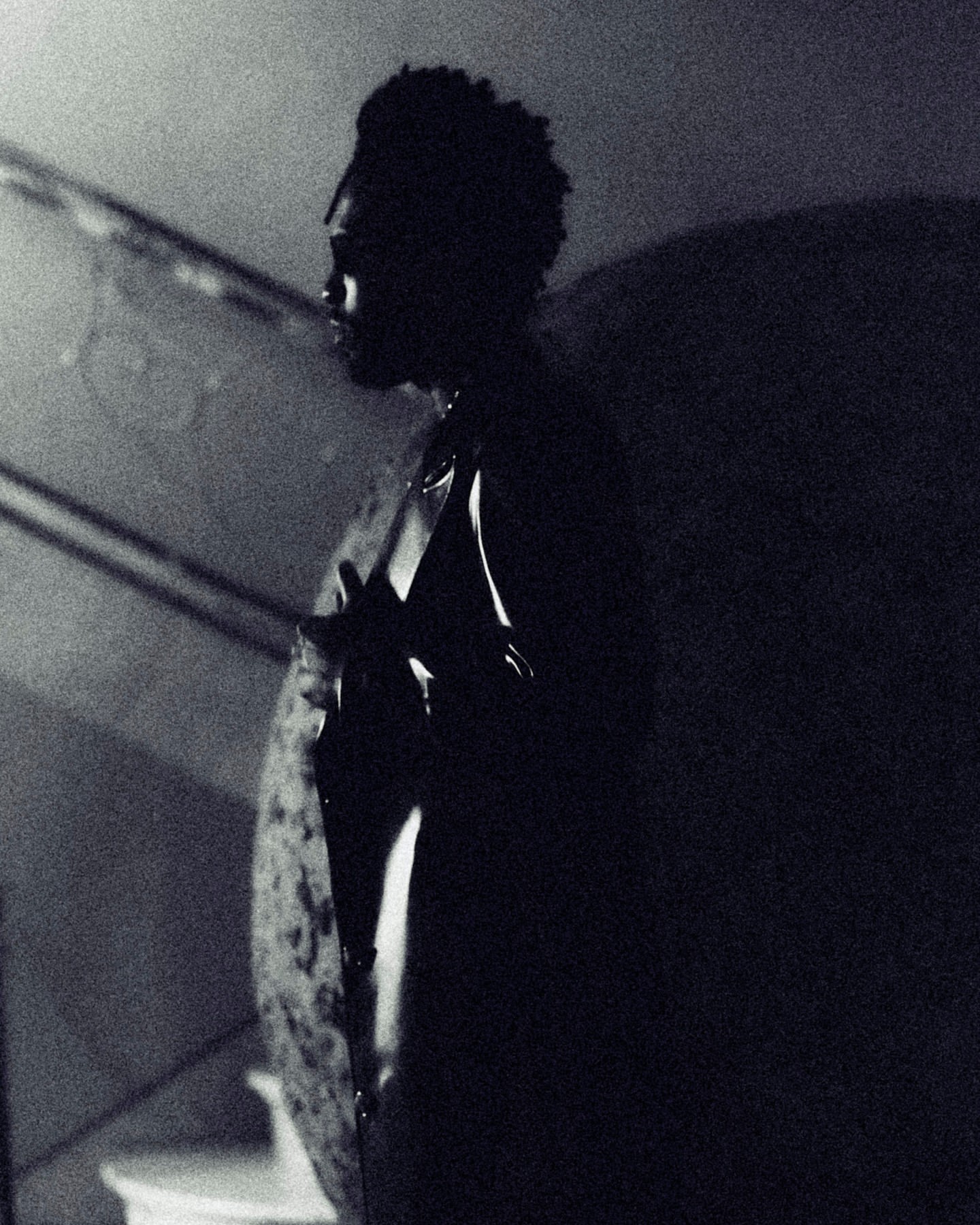 Monochrome portrait of an adult male in a shiny jacket against a shadowy background, stairs, and a wall.