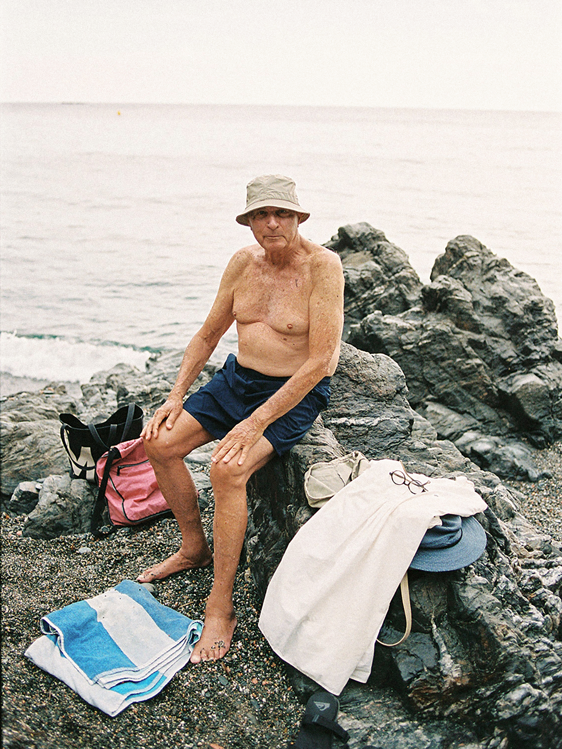 Elderly man in hat sits topless on rocky beach with a towel and beach bag. Ocean visible in the background.