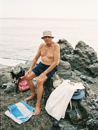 Elderly man in hat sits topless on rocky beach with a towel and beach bag. Ocean visible in the background.