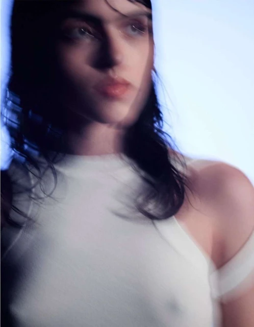 Close-up of a woman with wet, dark hair and light eyes, wearing a white top, against a light blue background.