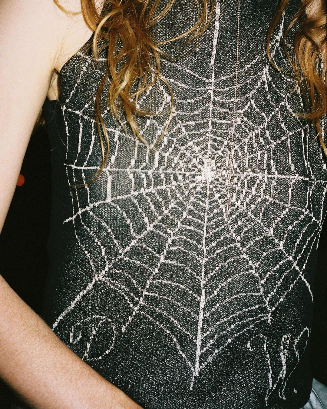 Close-up of a woman wearing a dark denim tank top with a white spiderweb design, her shoulder and arm visible.