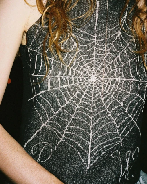 Close-up of a woman wearing a dark denim tank top with a white spiderweb design, her shoulder and arm visible.