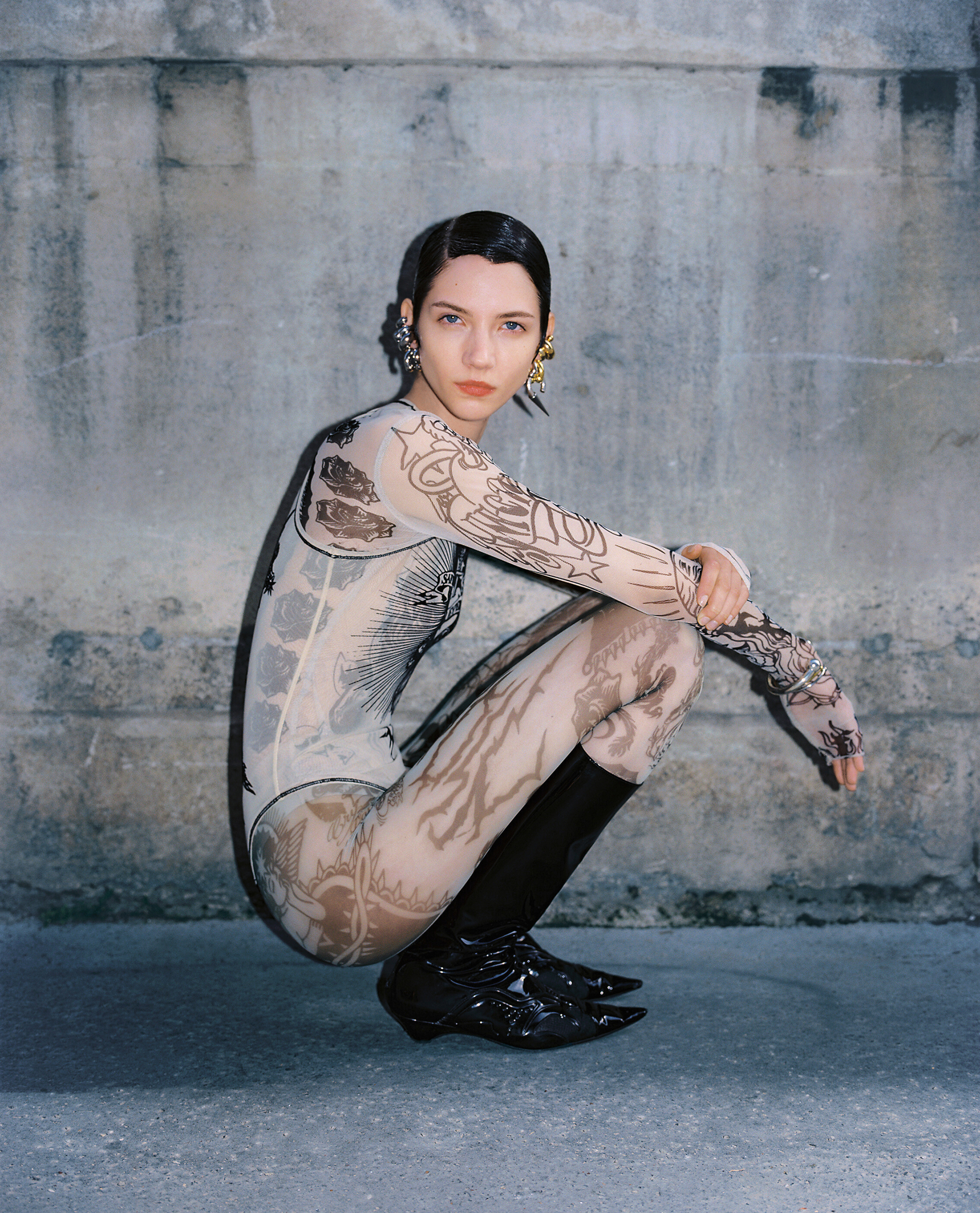 Woman wearing patterned bodysuit and black boots squats against a concrete wall. She wears large earrings and has a neutral facial expression.