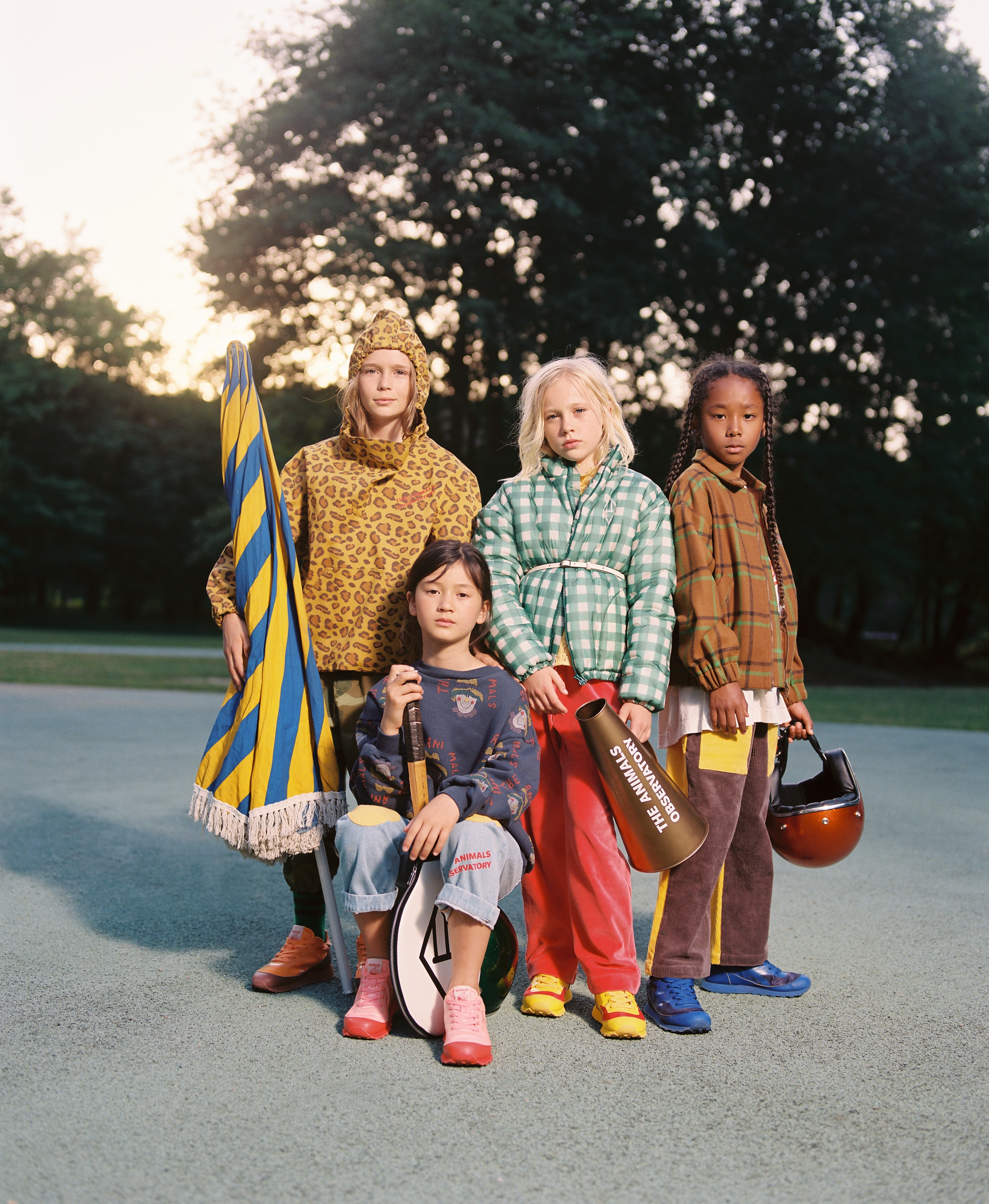 Four children in colorful vintage-style clothing pose outdoors in a park-like setting, holding props like a flag, tennis racket, and megaphone.