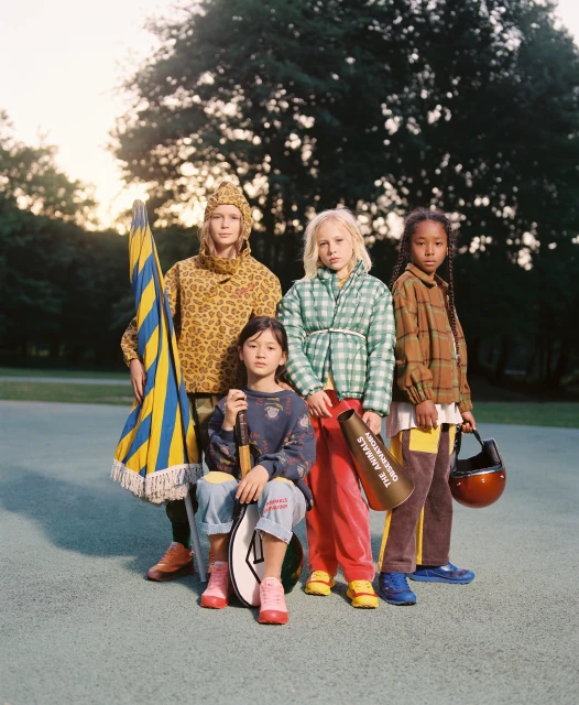 Four children in colorful vintage-style clothing pose outdoors in a park-like setting, holding props like a flag, tennis racket, and megaphone.