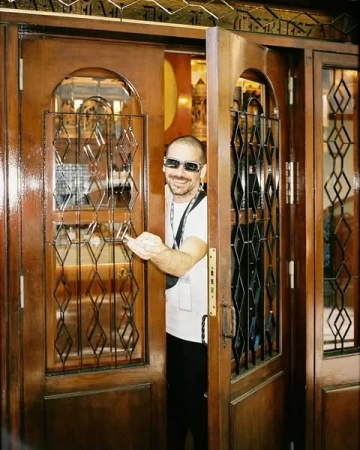 Man wearing sunglasses and a white t-shirt opening ornate wooden double doors with metal grilles