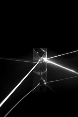 Prism refracts light, creating geometric patterns and illuminating a dark background with bright streaks in a black-and-white studio shot.