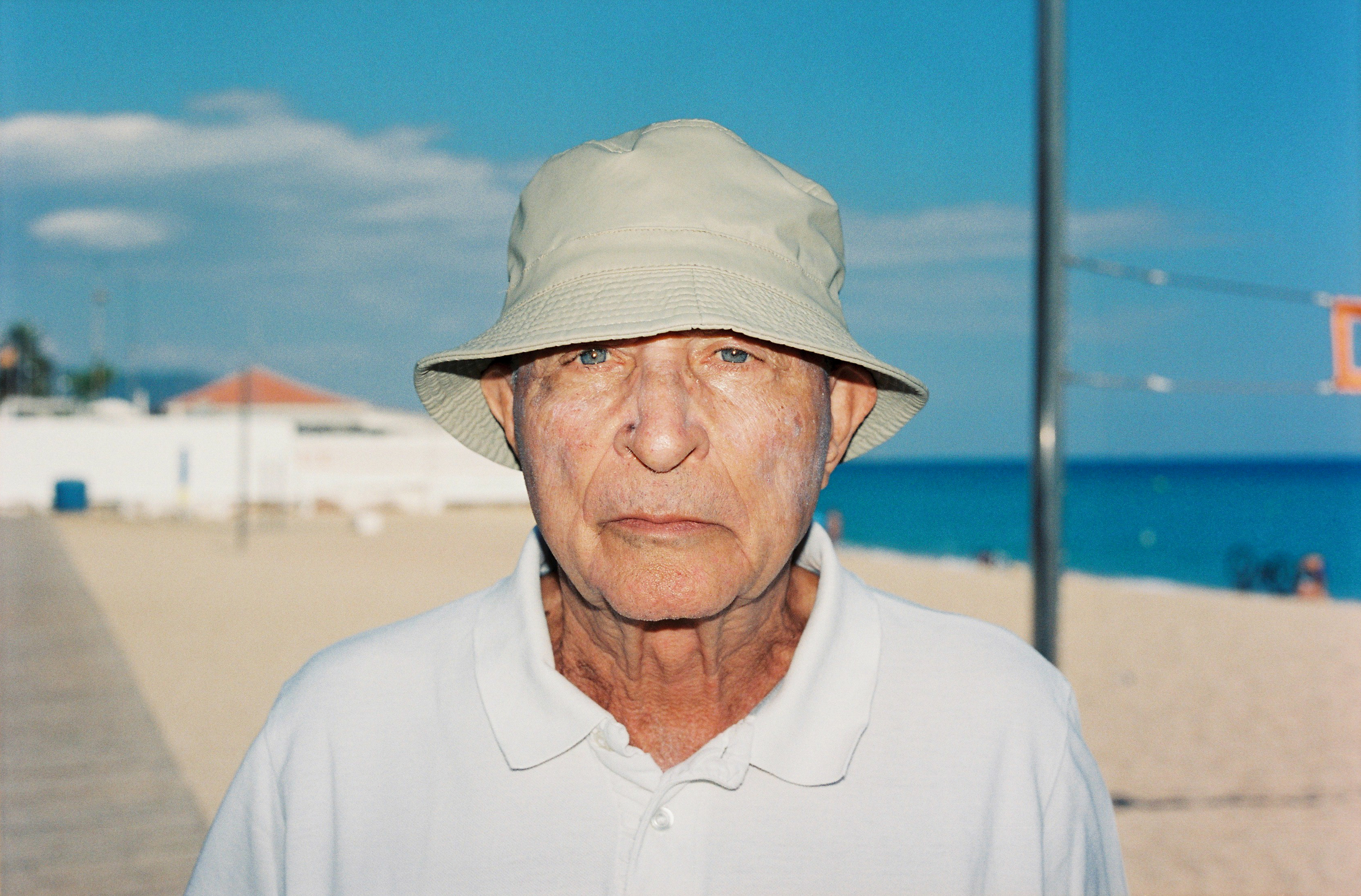 Portrait of an elderly man in a bucket hat at the beach on a sunny day, with ocean and a white building visible in the background.