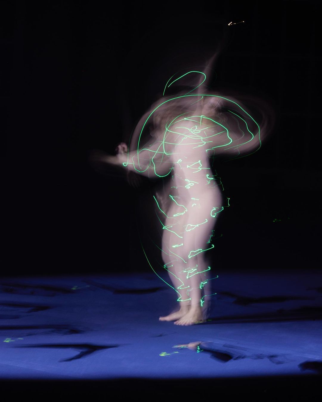 Blurred image of a nude woman in motion, illuminated by green light streaks, on a blue floor against a black background, long exposure photography.