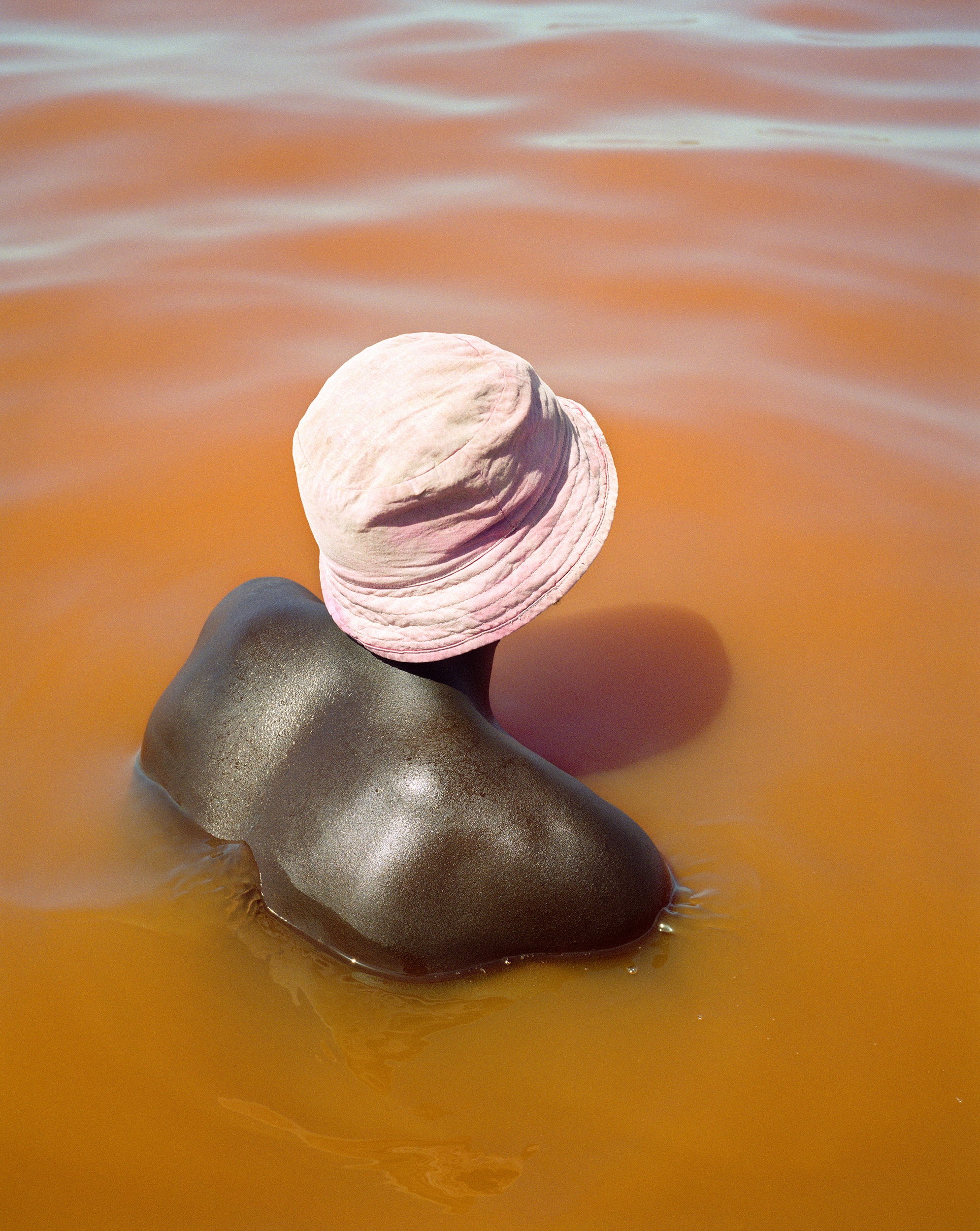 Dark-skinned adult stands waist-deep in orange water with a pink hat on, viewed from the back in a medium shot with soft lighting.