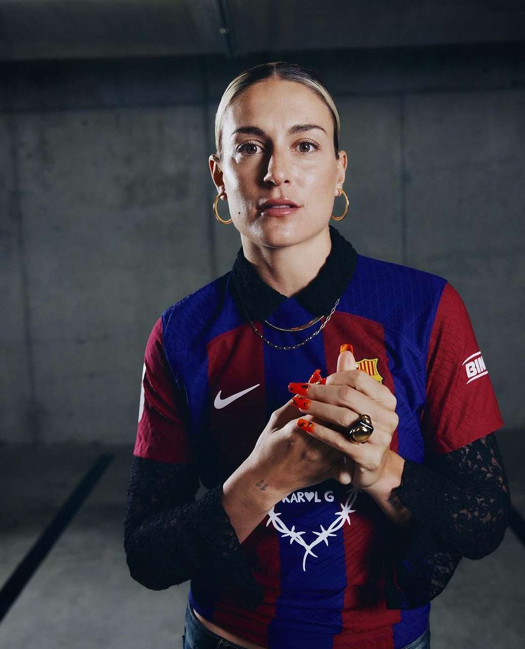 Portrait of a woman in a soccer jersey with a black lace undershirt, gold jewelry, and red nails, posing in a concrete setting.
