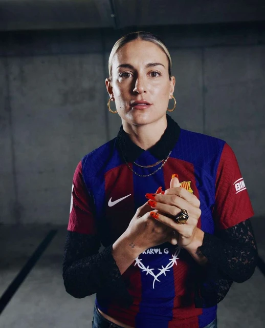 Portrait of a woman in a soccer jersey with a black lace undershirt, gold jewelry, and red nails, posing in a concrete setting.