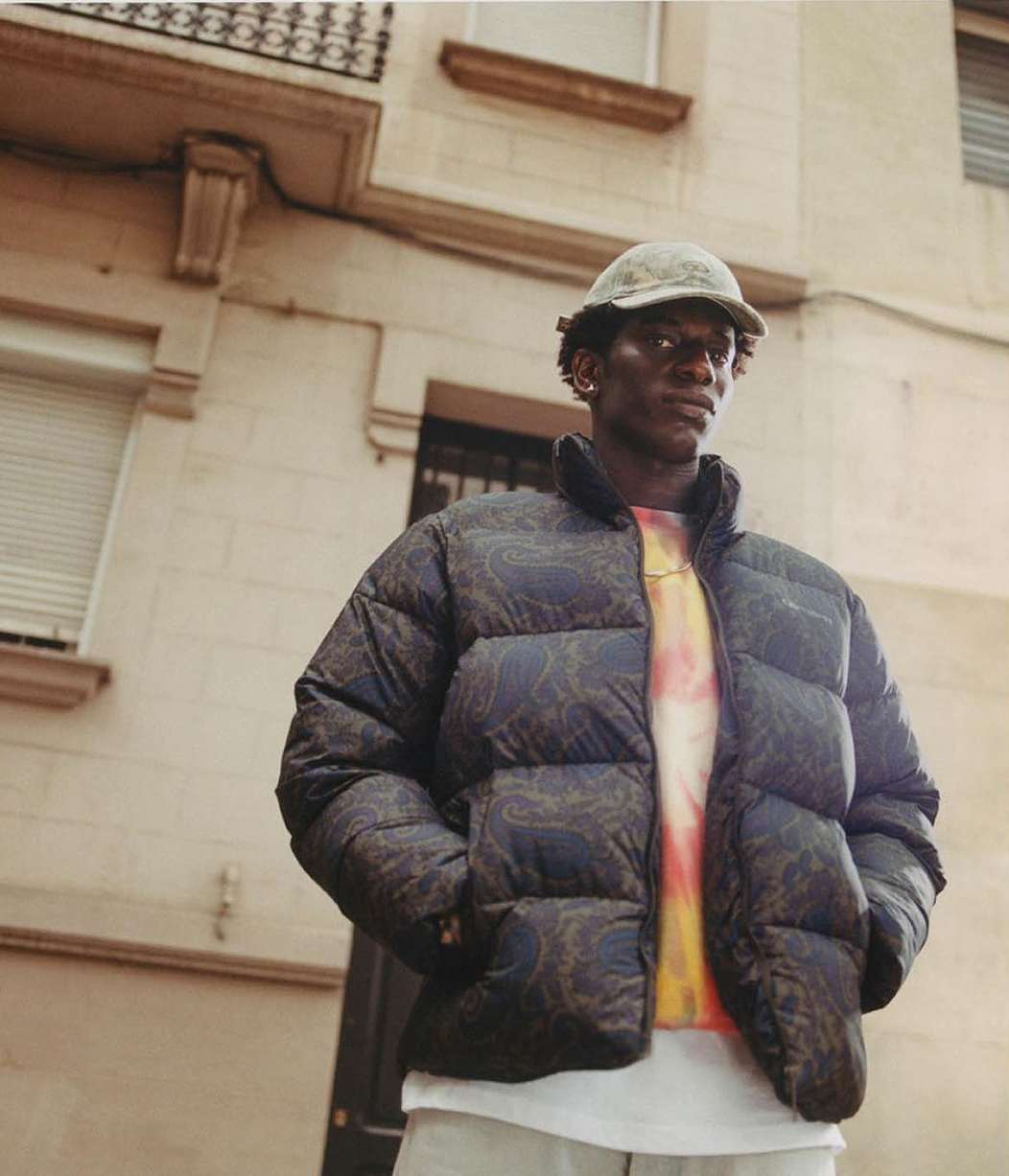 A stylish Black male model wears a patterned puffer jacket, cap, and tie-dye shirt against an urban building backdrop outdoors.