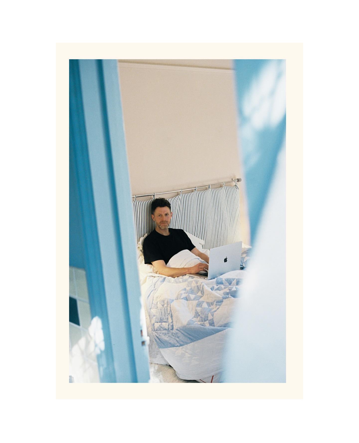 Man in bed using a laptop computer. The scene is framed by the blue doorway of the bedroom with a striped headboard and patterned duvet.