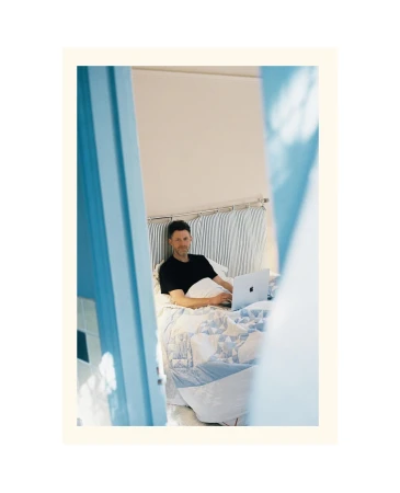 Man in bed using a laptop computer. The scene is framed by the blue doorway of the bedroom with a striped headboard and patterned duvet.