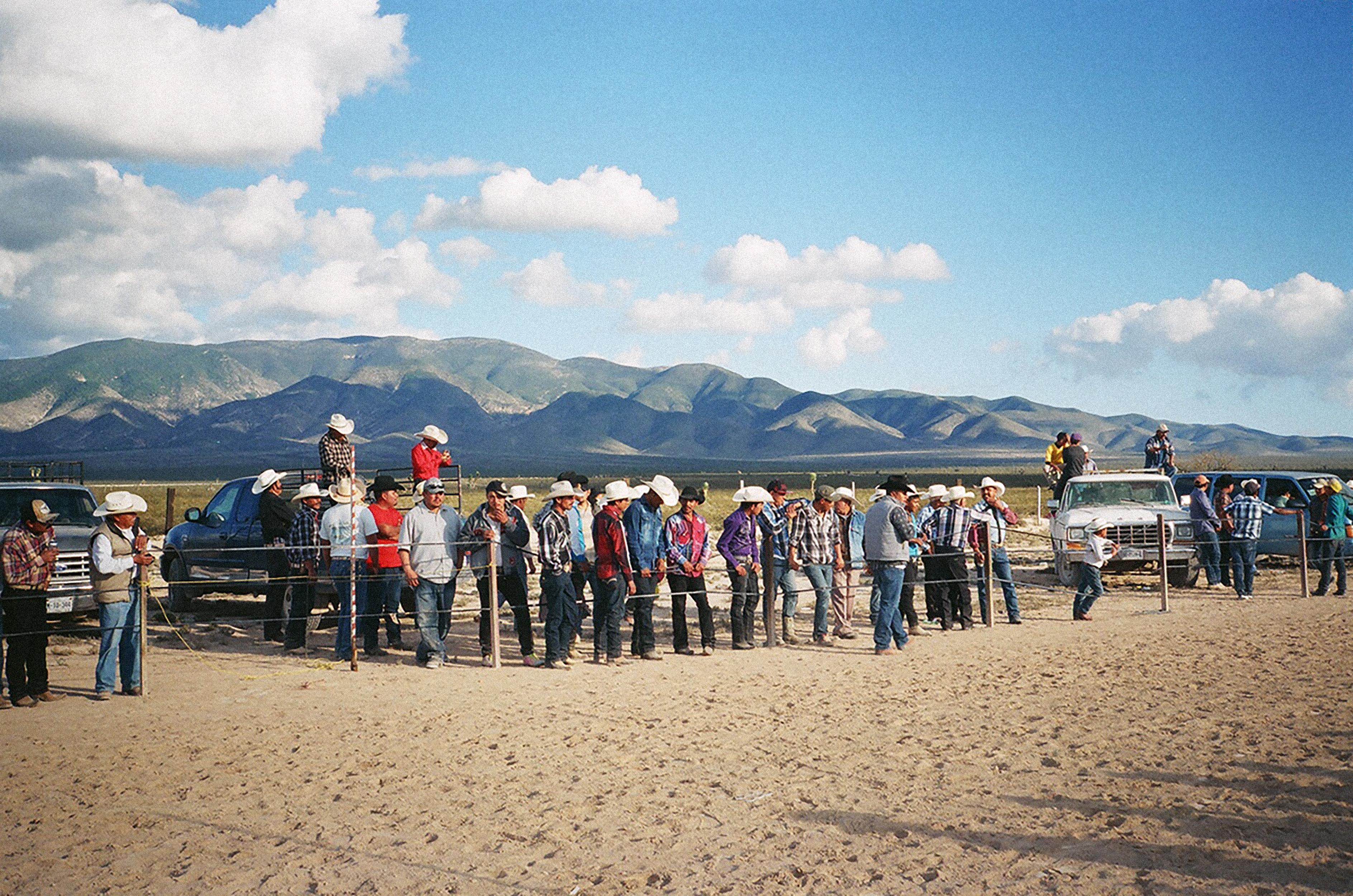 A group of people wearing cowboy hats standing on a dirt field, with trucks and mountains in the background under a cloudy sky.