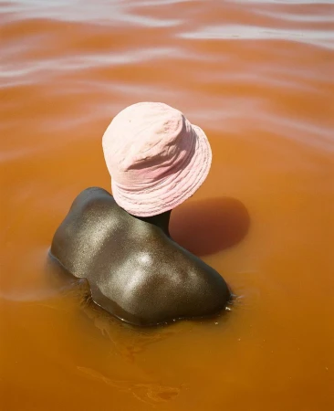 Person wearing a pink bucket hat submerged up to the shoulders in muddy orange water by photographer Dani Pujalte