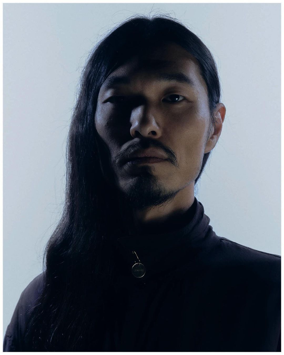 Portrait of a man with long dark hair and facial hair wearing a dark zip-up jacket against a plain light background